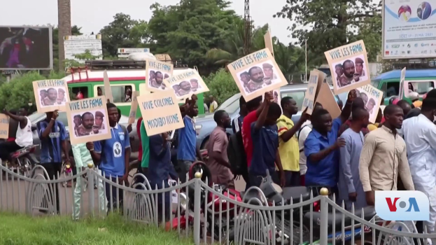 Eine Gruppe von Menschen h├Ąlt Schilder, wahrscheinlich protestierend, vor einem Zaun mit Fahrzeugen, B├Ąumen, Pf├Âhlen, Absperrungen und einem Bildschirm im Hintergrund, unter einem sichtbaren Himmel mit Gras am unteren Bildrand.