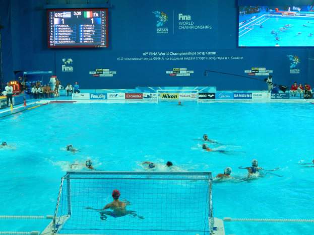 Eine Gruppe von Menschen, die Wasserball in einem große Pool mit einem Netz in der Mitte spielen, mit Zuschauern auf der linken Seite und einem Bildschirm mit Text und Logos im Hintergrund bei den FINA-Weltmeisterschaften 2016 in Kazan.
