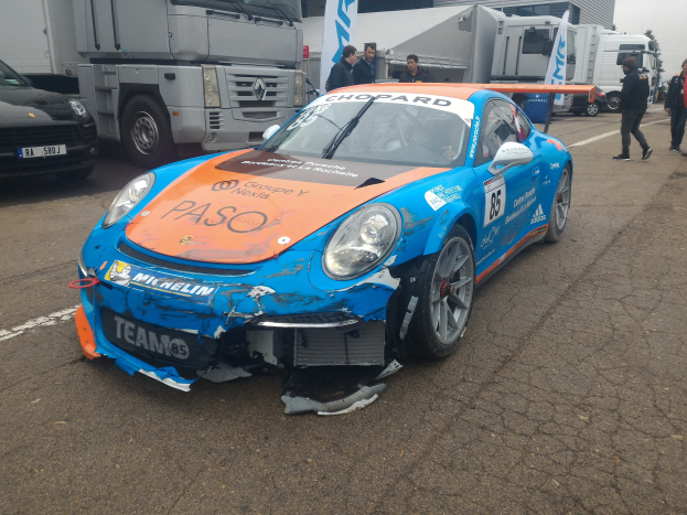 Blauer und oranger Porsche 911 GT3 RSR krachte in einen Lastwagen am Straßenrand mit Menschen in der Nähe, vor Häusern, Bäumen und einem klaren blauen Himmel.