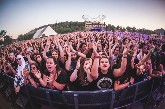 A large crowd at a music festival raises their hands in front of a railing, with trees, buildings, and a clear blue sky in the background.