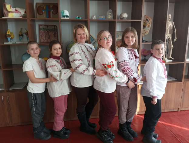 Eine Gruppe von Kindern in traditioneller ukrainischer Kleidung vor einem Bücherregal mit Spielzeugen und anderen Gegenständen, was auf eine ukrainische Schulumgebung hinweist.