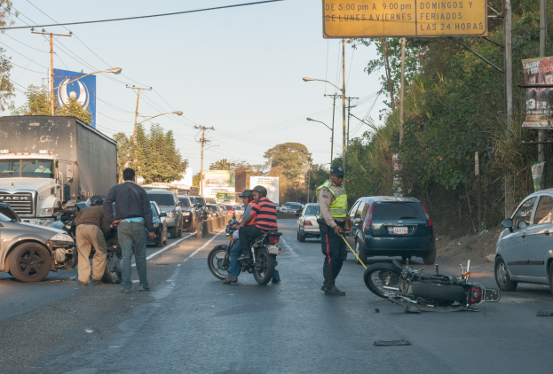 Eine Gruppe von Menschen steht um ein verunglücktes Motorrad auf der Stra√enseite mit mehreren Fahrzeugen, darunter ein Lastwagen, und Hintergrundelementen wie Bäumen, Masten, Lichtern, Schildern und dem Himmel.