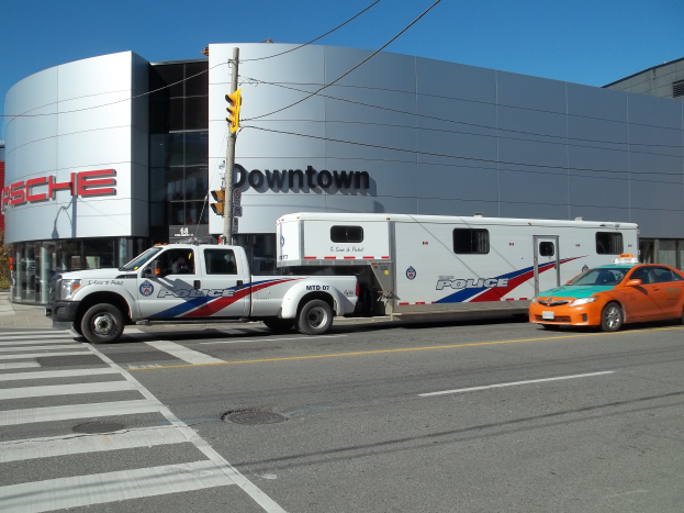 Ein Polizei-Lkw vor einem Gebäude mit Text, einer Ampel mit Mast und Kabeln im Vordergrund und Bäumen mit einem klaren blauen Himmel im Hintergrund.