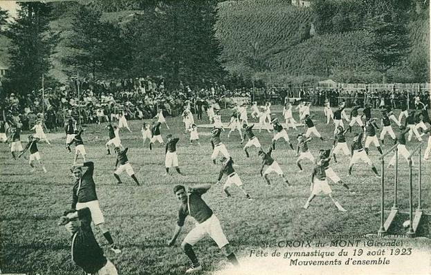 Schwarzes und weißes Foto einer Gruppe von Menschen, die auf einem grasbewachsenen Feld mit Bäumen, Gebäuden und einem klaren Himmel Fußball spielen, datiert "Fête de Gymnastique du 19 août 1923".