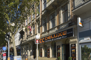 Eine belebte Stadtstraße mit verschiedenen Gebäuden, Fahrzeugen, Fußgängern und einem prominenten Gebäude in der Mitte mit einer Tafel für "CC Deluxe Cafe & Cocktail Bar" unter einem klaren blauen Himmel.