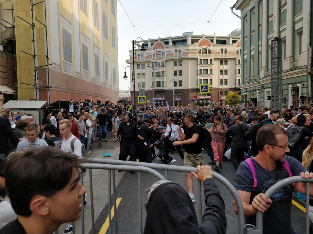 A large crowd of people stands behind a metal barricade on a road, with buildings, trees, and light poles in the background, some holding cameras during a protest.