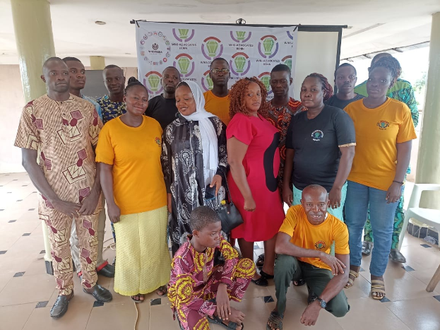 Group of people posing for a photo in front of a banner reading "WWA Advocates in Kenya" with chairs, pillars, a wall, and ceiling lights in the background.