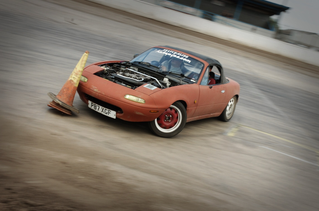 Eine Person, die ein rotes Auto auf einer Rennstrecke fährt und auf eine Verkehrsinsel zusteuert, mit einem unscharfen Hintergrund, der hohe Geschwindigkeit andeutet.