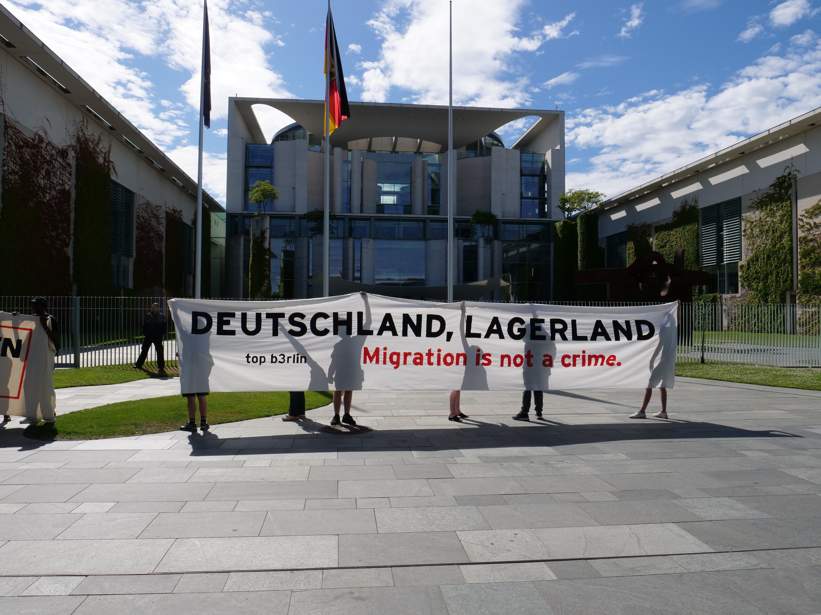 Eine Gruppe von Menschen mit einem Transparent, auf dem "Deutschland, Lagerland Migration ist kein Verbrechen" steht, vor einem Gebäude mit einem Metallzaun, umgeben von Gras, Pflanzen, Bäumen und Fahnen auf Ständen unter einem bewölktem Himmel.
