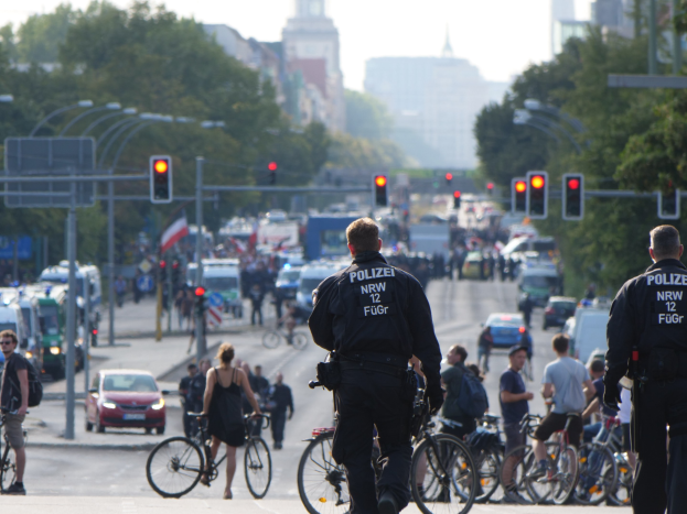 Zwei Polizeibeamte neben einer Gruppe von Radfahrern auf einer Straße mit Fahrzeugen, Verkehrszeichen, Bäumen, Gebäuden und einem klaren Himmel im Hintergrund.
