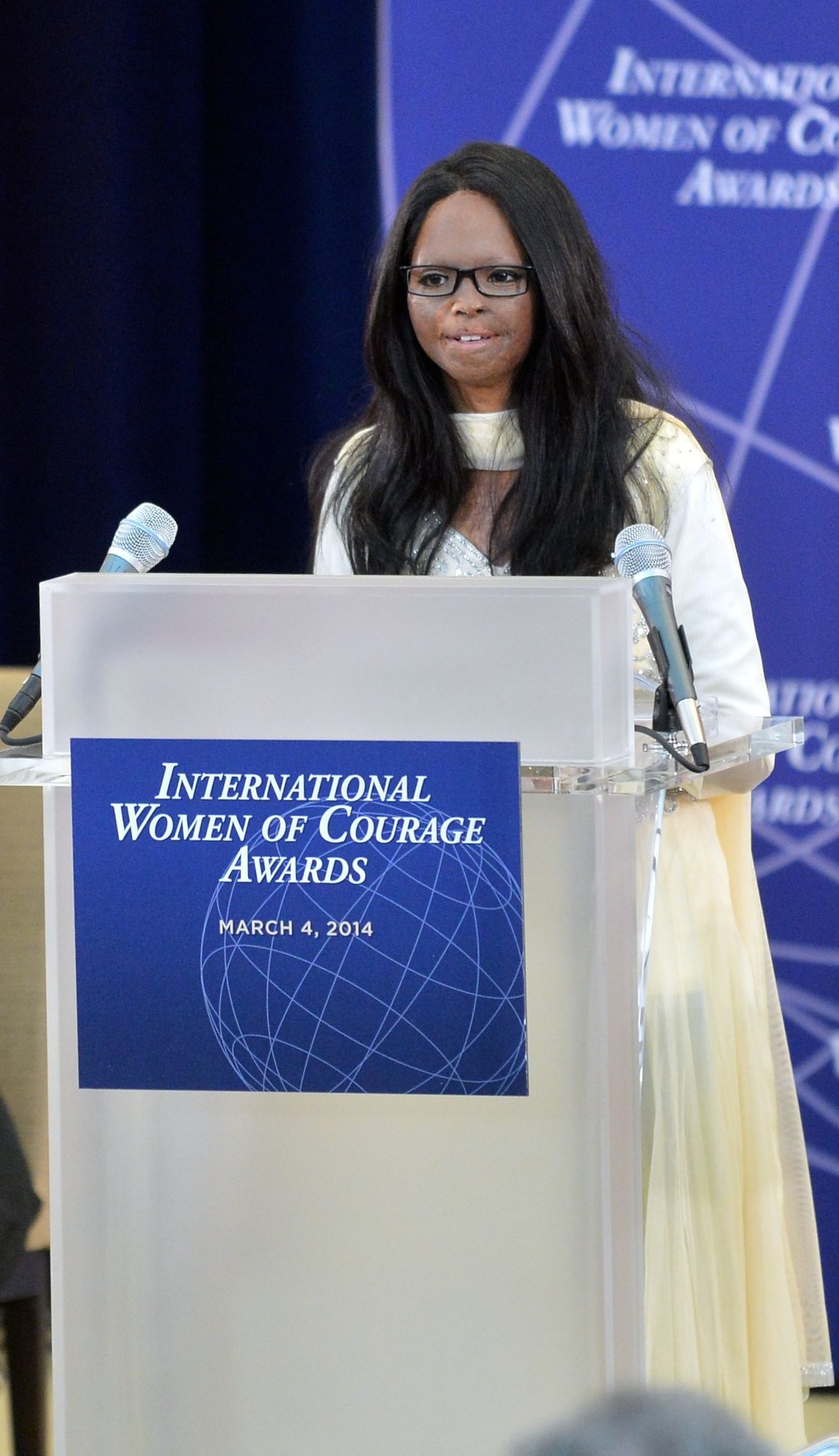Woman in a white dress and spectacles speaking at a podium with two microphones and a board, in front of a crowd, with a blue banner labeled "International Women of Courage Awards 2014" in the background.