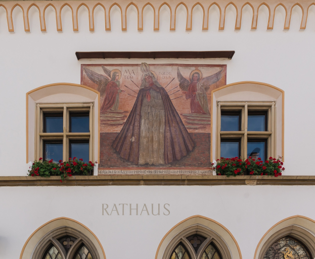 Building with windows and a mural on the side, surrounded by flowering plants, with the word "Rathaus" at the bottom.