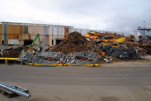 Ein großer Haufen Schrott vor einem Gebäude, umgeben von verschiedenen Metallobjekten auf der Straße, mit Bäumen, Masten und einem klaren blauen Himmel im Hintergrund.
