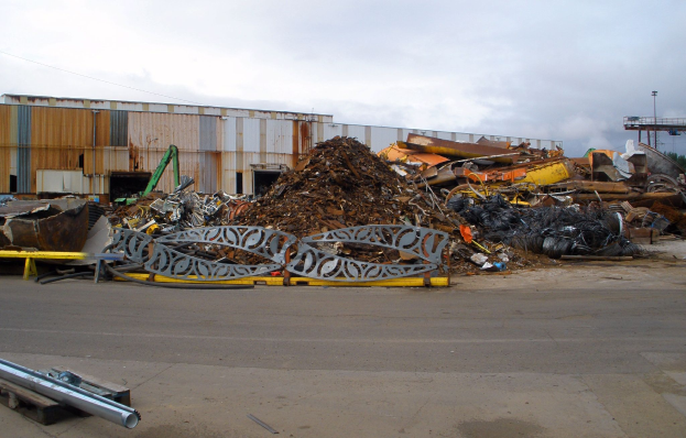Ein großer Haufen Schrott vor einem Gebäude, umgeben von verschiedenen Metallobjekten auf der Straße, mit Bäumen, Masten und einem klaren blauen Himmel im Hintergrund.