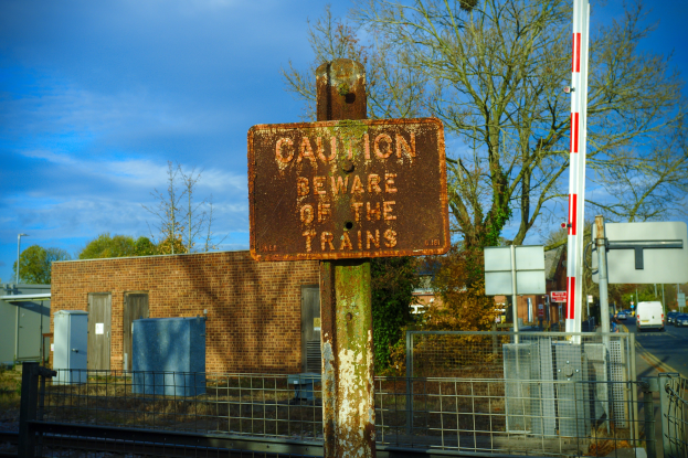 Vorsichtsschild auf einem Bahngleis, umgeben von einem Metallzaun, Bäumen, Strommasten, einem Gebäude mit Fenstern, Containern, einem Straßenmast, Fahrzeugen auf der Straße und einem bewölkten Himmel.