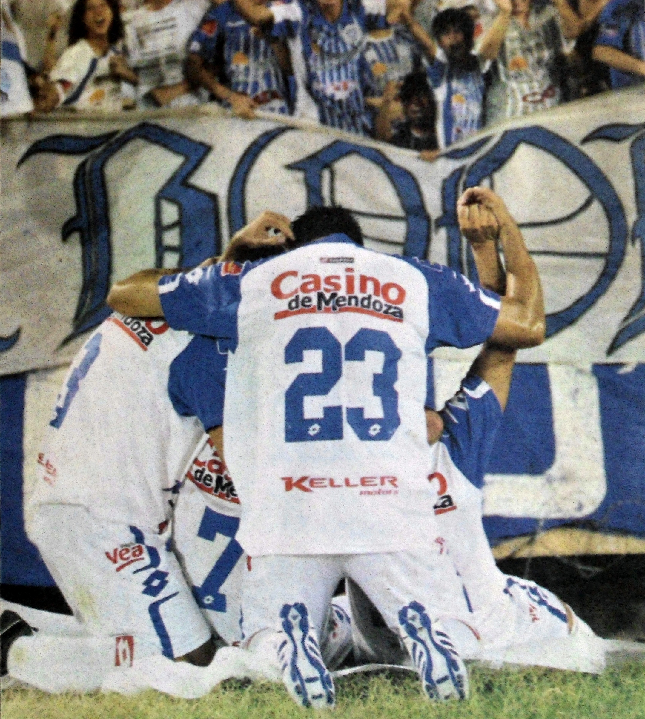 Eine Gruppe von Fußballspieler feiert ein Tor auf dem Boden vor einer jubelnden Menge, mit einem Banner mit Text im Hintergrund.