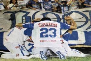 Eine Gruppe von Fußballspieler feiert ein Tor auf dem Boden vor einer jubelnden Menge, mit einem Banner mit Text im Hintergrund.