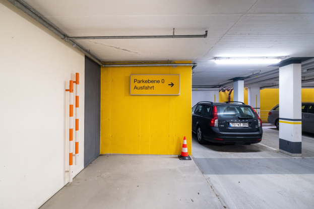Eine Tiefgarage mit zwei geparkten Autos, einem Verkehrskegel auf dem Boden, einer wandmontierten Texttafel, Deckenleuchten und einem sichtbaren Rad.