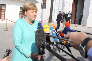 Bundeskanzlerin Angela Merkel spricht vor einem Gebäude mit Fenstern und einer Tür zu den Medien, umgeben von einer Gruppe von Menschen, mit einer Kamera und einem Mobiltelefon auf der rechten Seite und einigen Pfählen mit daran gebundenen Seilen.
