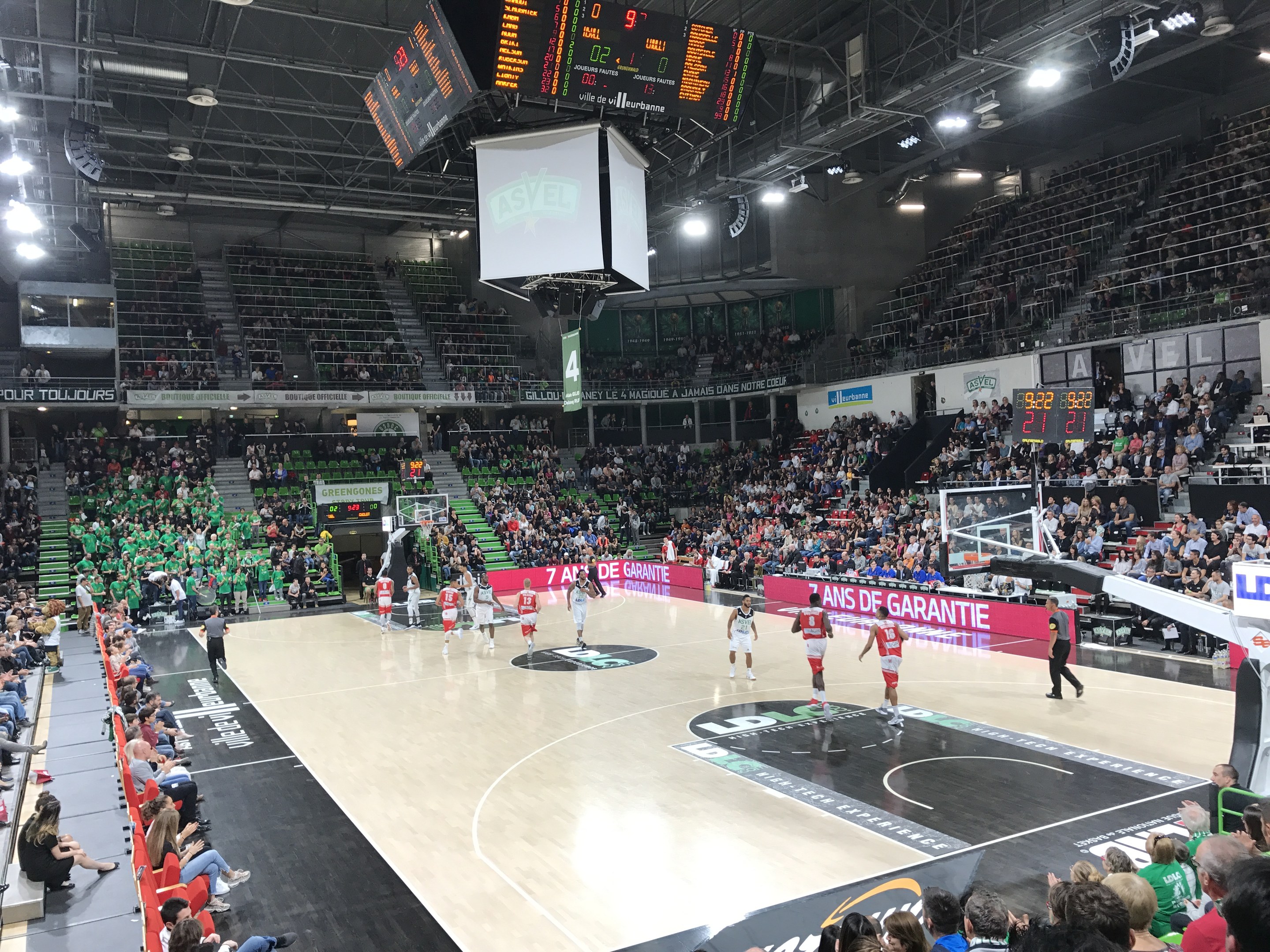 Basketballspiel in einer großen Arena mit Zuschauern, mit einem Scoreboard, das "Asvel vs Galatasaray" anzeigt, während ein Spiel zwischen den beiden Mannschaften stattfindet.