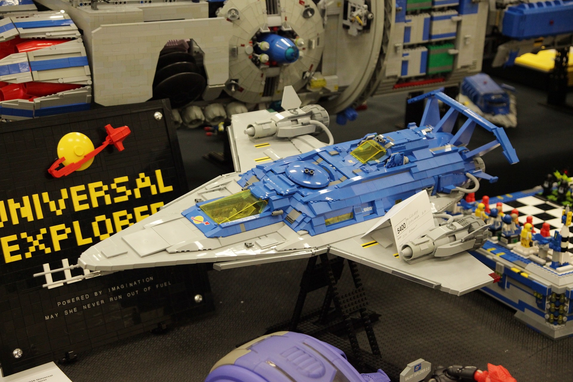 Lego-Modell eines Raumschiffs auf einer Convention ausgestellt, umgeben von Spielzeugen und einer Tafel mit Text.