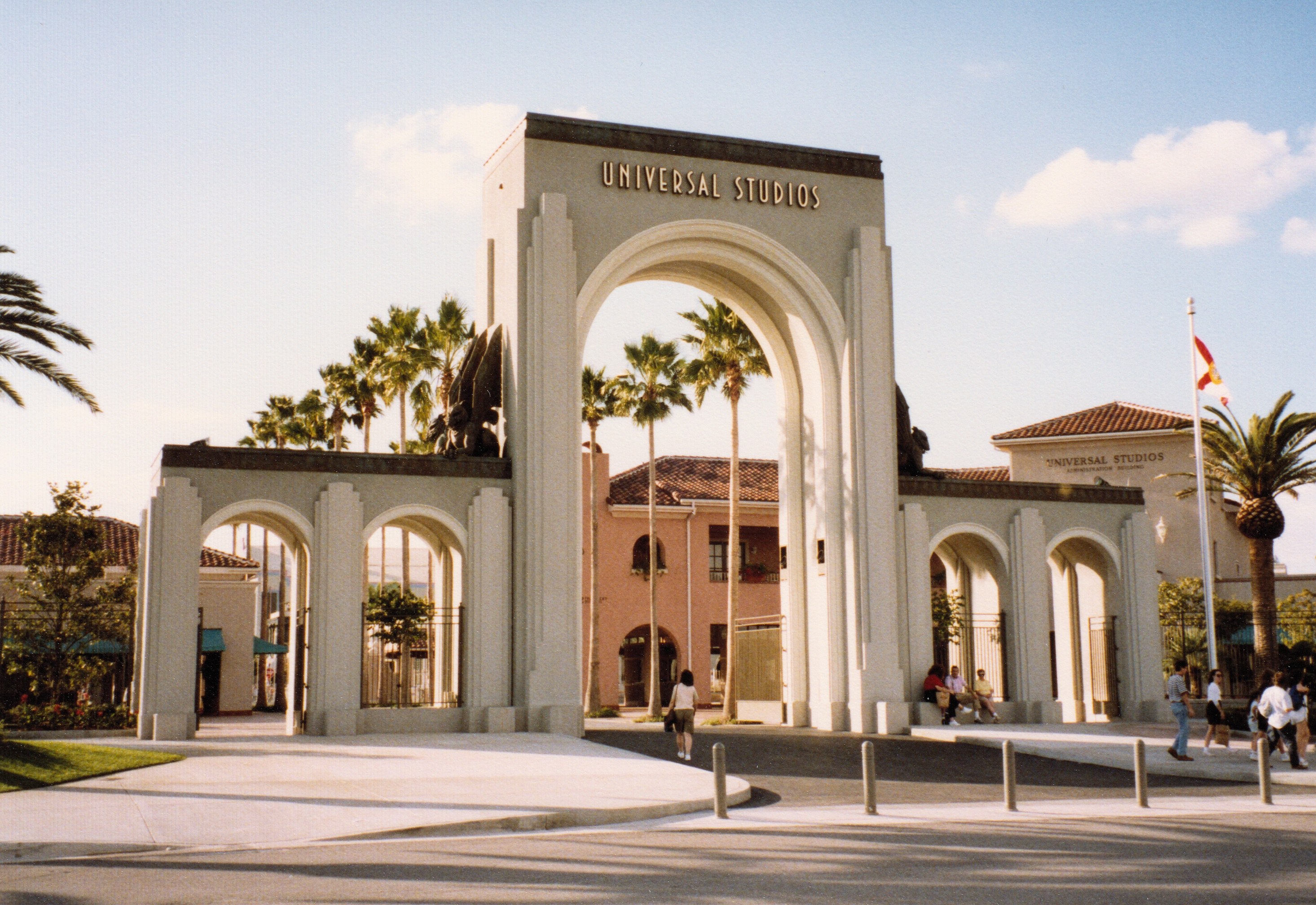 Eingang zum Universal Studios Hollywood mit einem Bogen mit Text, einer Statue, Menschen, Bäumen, Gebäuden, einem Fahnenmast mit einer Fahne.
