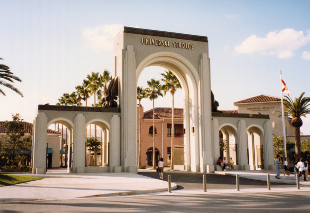 Eingang zum Universal Studios Hollywood mit einem Bogen mit Text, einer Statue, Menschen, Bäumen, Gebäuden, einem Fahnenmast mit einer Fahne.