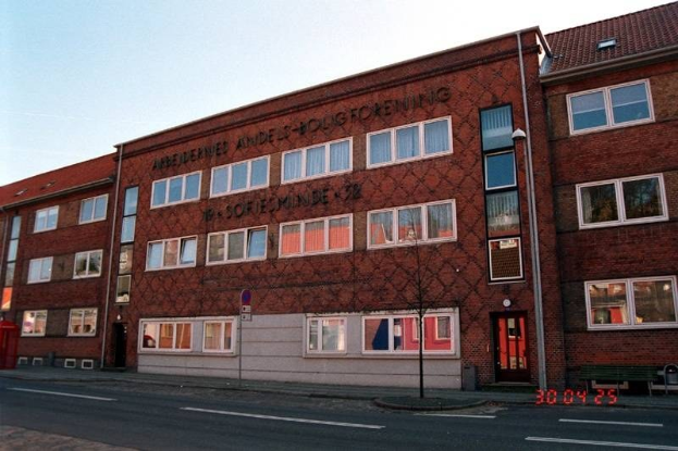 Großes Backsteingebäude mit vielen Fenstern, als deutsche Bundespolizei-Hauptquartier bezeichnet, auf einer Straße mit einem Gehweg, einem Baum, einer Bank, einem Laternenpfahl und einem roten Briefkasten, unter einem sichtbaren Himmel.
