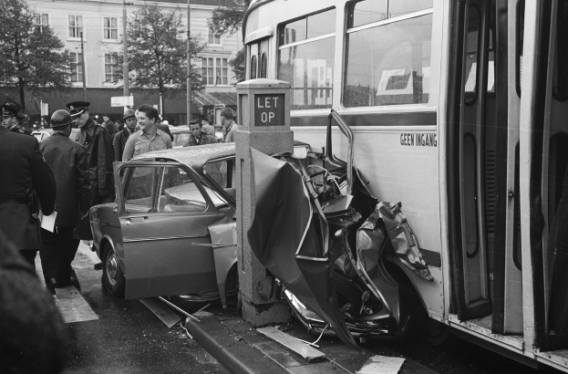 Schwarz-weißes Bild eines Busses, der auf der Seite der Straße in ein Auto gekracht ist, mit Menschen und Bäumen im Hintergrund.