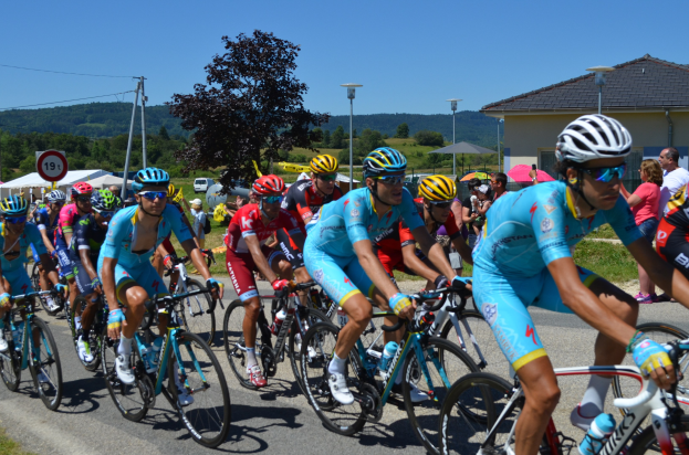 Gruppe von Radfahrern in Sportkleidung auf einer Stra├če mit Gras an beiden Seiten, umgeben von Zelten, Fahrzeugen, B├Ąumen und Geb├Ąuden unter einem klaren blauen Himmel.