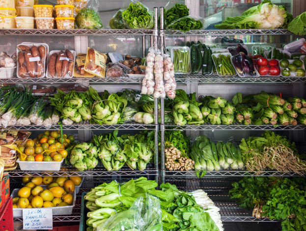 Ein Gang in einem Supermarkt mit frischem Obst und Gemüse in Plastikverpackungen, Kisten und einer Texttafel sowie einem Glasfenster im Hintergrund.