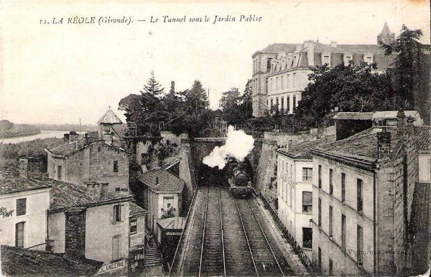 Black and white photo of a train on tracks passing buildings, trees, and water with text "la réole gironde - le tunnel sous le jardin public" at the top.