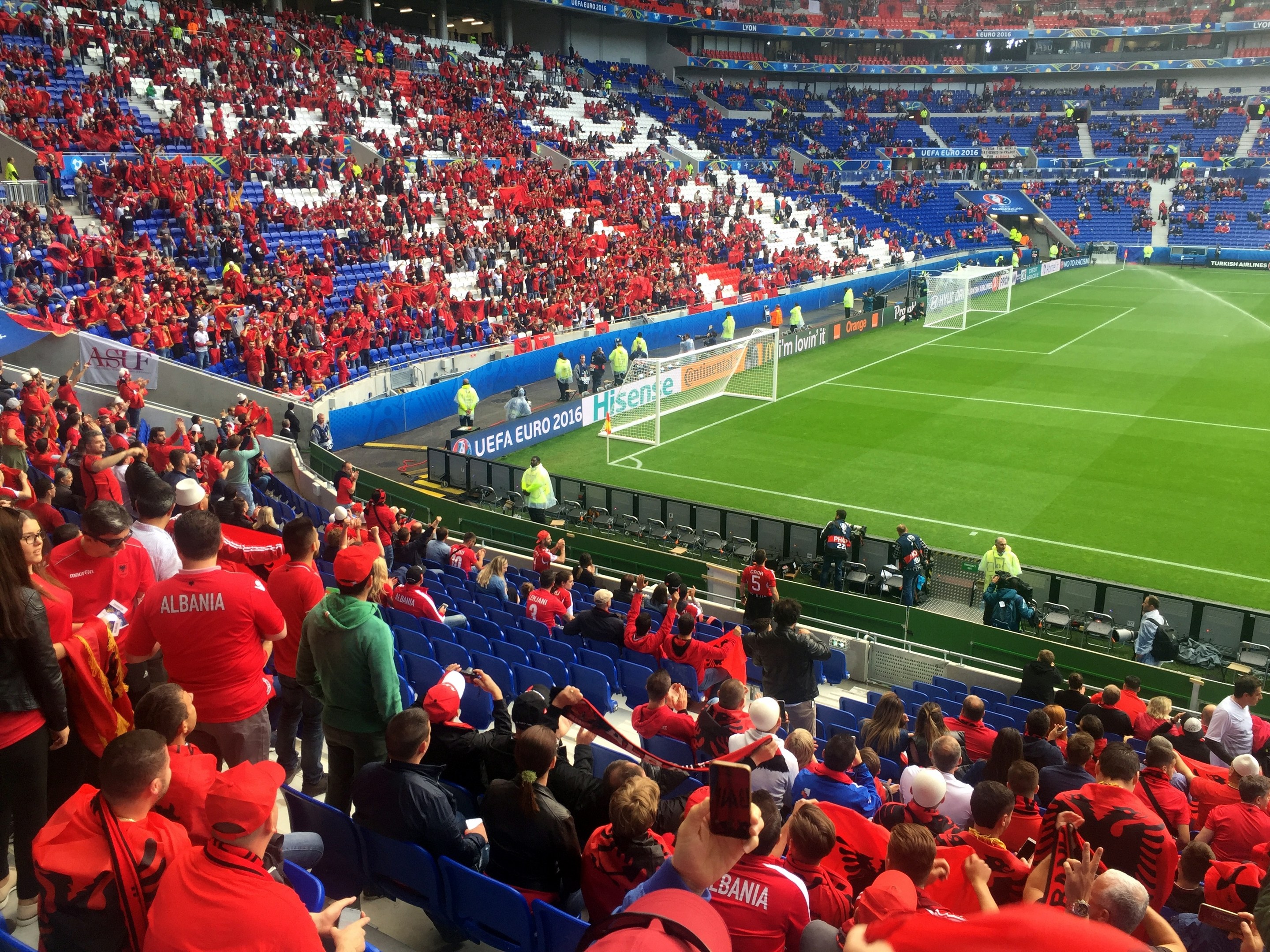 Eine große Menschenmenge in einem Stadion schaut ein Fußballspiel, mit Sitzplätzen, Stehplätzen, Bannern, einem Torpfosten und Deckenbeleuchtung.