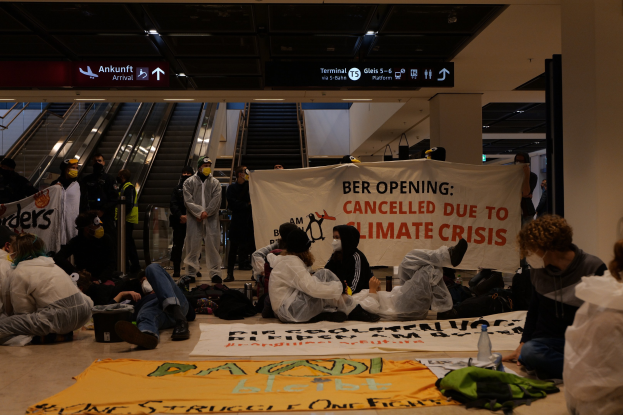 People sitting on an airport floor holding a "Berlin Cancelled Due to Climate Crisis" banner with scattered belongings and escalators in the background.