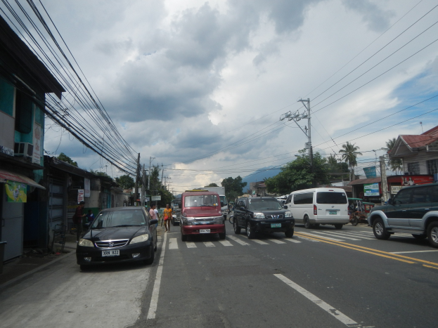 Stadtstraße mit parkenden Autos, Fußgängern auf dem Gehweg, Gebäuden, Strommasten mit Drähten, Bäumen und einem bewölkten Himmel.
