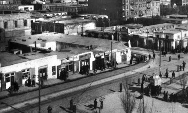 Schwarzes und weißes Foto einer Stadtstraße mit Fußgängern, Gebäuden, Bäumen, Strommasten und Drähten, die die historische städtische Umgebung widerspiegeln.