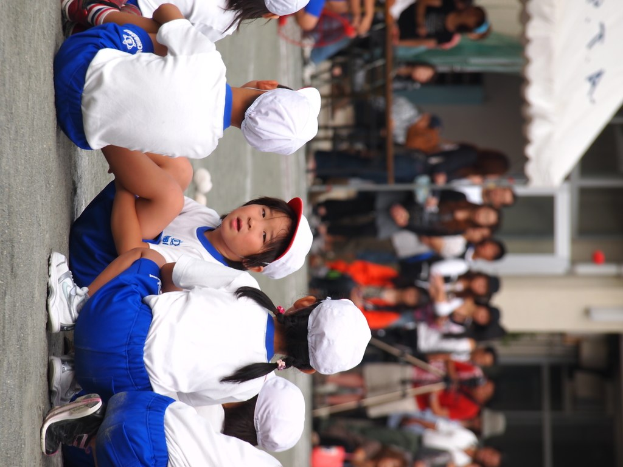 Kinder sitzen auf der Straße mit Menschen im Hintergrund, ein Stativständer, ein Zelt oben und Glaswände eines Gebäudes auf der rechten Seite.