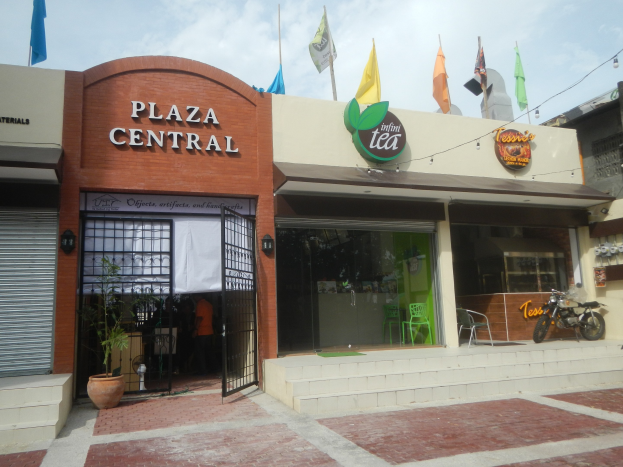A central plaza in Cebu City, Philippines, with buildings, motorbikes, outdoor seating, houseplants, flags, signage, overhead electric lights and cables, and a cloudy sky.