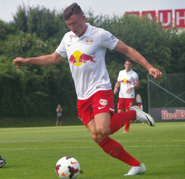 Professioneller Fussballspieler, der einen Ball auf einem grünen Feld mit Bäumen und einem klaren blauen Himmel im Hintergrund tritt, RB Leipzig-Logo sichtbar.