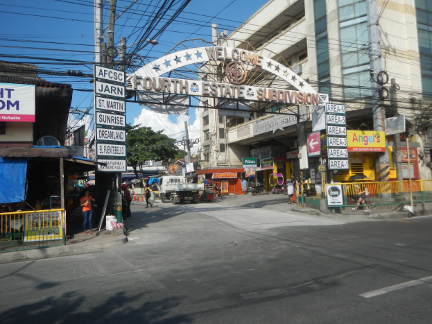 Eine belebte Stadtstraße mit fahrenden Fahrzeugen, Fußgängern auf dem Gehweg, einem Torbogen mit Text, Gebäuden mit Fenstern, Strommasten und -leitungen, Bäumen und einem bewölkten Himmel, mit einem "Fourth Estate Subdivision"-Straßenschild in der Mitte.