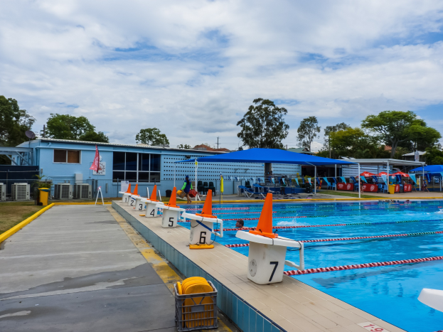 Großes Freibad mit schwimmenden Menschen, umgeben von Bahntrennern, Verkehrskegeln, Stühlen, Sonnenschirmen, einem Gebäude mit Fenstern, einer Flagge, Bäumen und einem bewölkten Himmel.