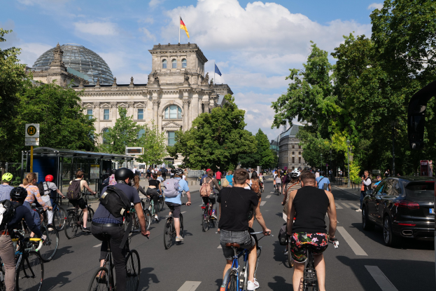 Gruppe von Menschen, die auf Fahrrädern eine von Bäumen gesäumte Straße in Berlin, Deutschland, entlangfahren, mit Gebäuden und einem Bushaltestellen-Schild im Hintergrund, unter einem bewölkten Himmel und einer Flagge, die auf einem Gebäude weht.