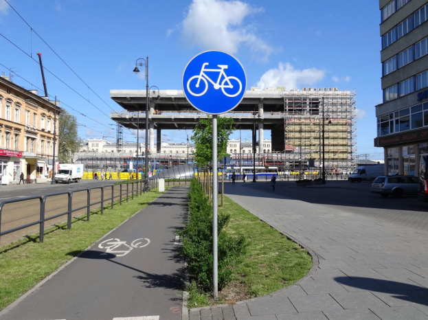 Blaues Verkehrszeichen mit der Aufschrift "Radweg in Berlin" auf einem Pfahl neben einem Gebäude mit Gras, Bäumen und Fahrzeugen im Hintergrund unter einem bewölktem Himmel.