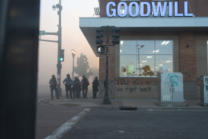 Eine Gruppe von Polizisten in Helmen und mit Waffen in der Hand steht vor einem Goodwill-Laden mit Glasw├Ąnden, mit Laternenmasten, Verkehrsampeln, B├Ąumen und einem klaren Himmel im Hintergrund.