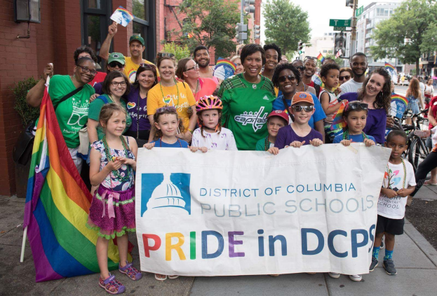 Gruppe von Menschen mit einem Banner 'District of Columbia Public Schools Pride in DCPS' vor einem Gebäude mit Bäumen, Straßenlaternen und einem bewölkten Himmel.