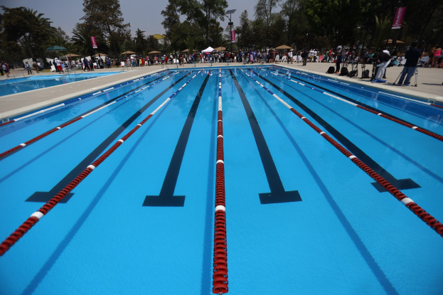 Großer Schwimmbad mit Bahnentrennern, umgeben von Menschen mit Taschen und Gegenständen, mit Zelten, Transparenten, Masten, einem Schild und einem bewölkten Himmel im Hintergrund.