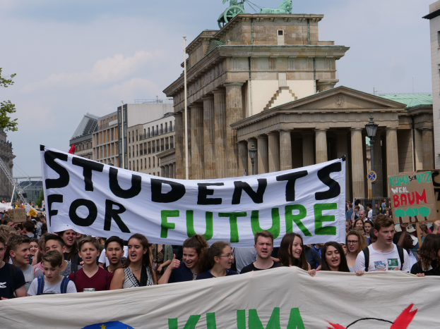 Gruppe von Schülern marschiert in Berlin mit buntem "Students for Future"-Schild gegen Gebäude, Bäume und Himmel.