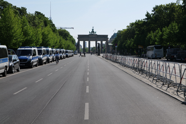 Lange Reihe von Polizeiwagen am Straßenrand vor dem Brandenburger Tor in Berlin, Deutschland, mit Menschen auf Fahrrädern und Fußgängern, Absperrungen, Bäumen, einem Bogen mit Statuen im Hintergrund und sichtbarem Himmel.