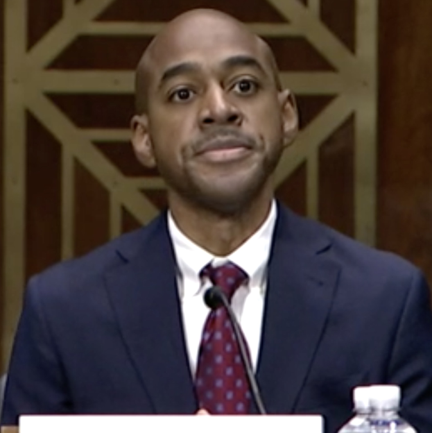 A suited man testifying before the Senate Judiciary Committee at a table with a nameplate, microphone, and water bottle, facing a wall.
