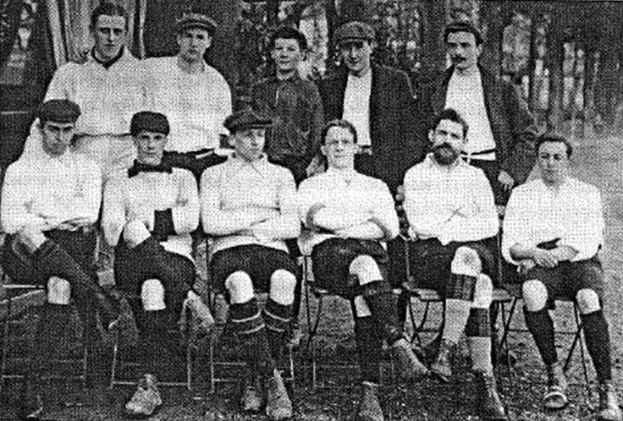 Schwarzes und wei%C3%9Fes Mannschaftsfoto des West Bromwich Albion Football Club aus dem Jahr 1918, das sitzende und stehende M%C3%A4nner mit B%C3%A4umen im Hintergrund zeigt.